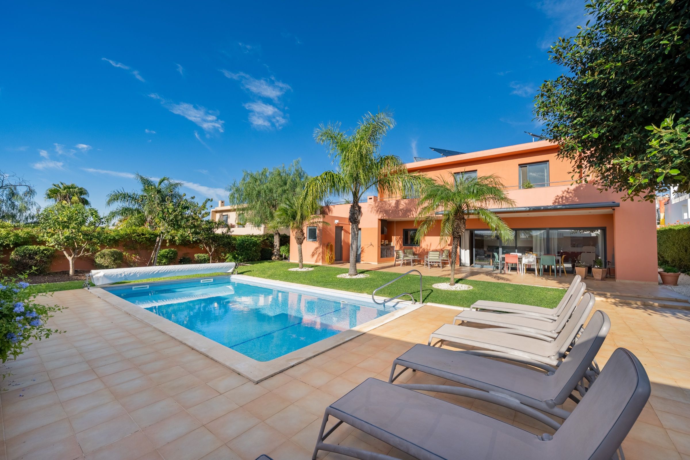 Pool of a pink villa, with a lawn, palm trees and sun loungers on a clear blue day