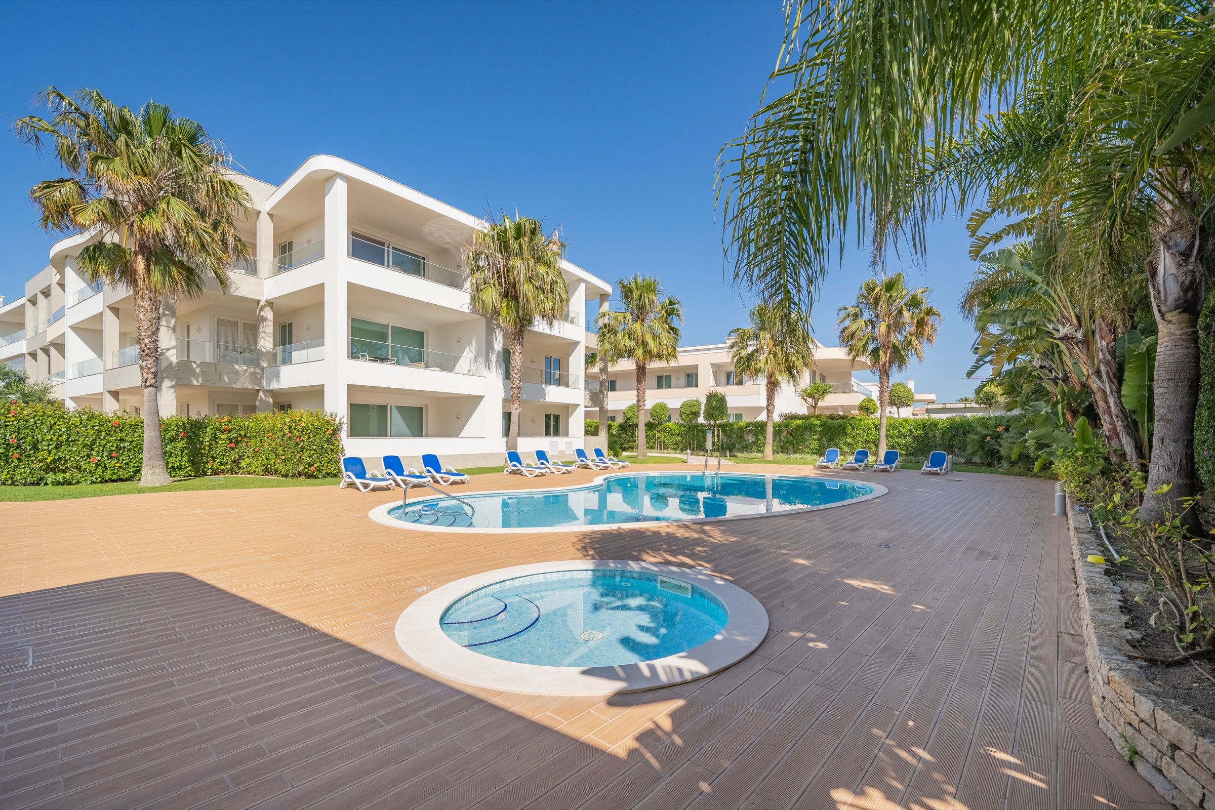 White apartment complex with a pool, hot tub, palm trees and blue sun loungers