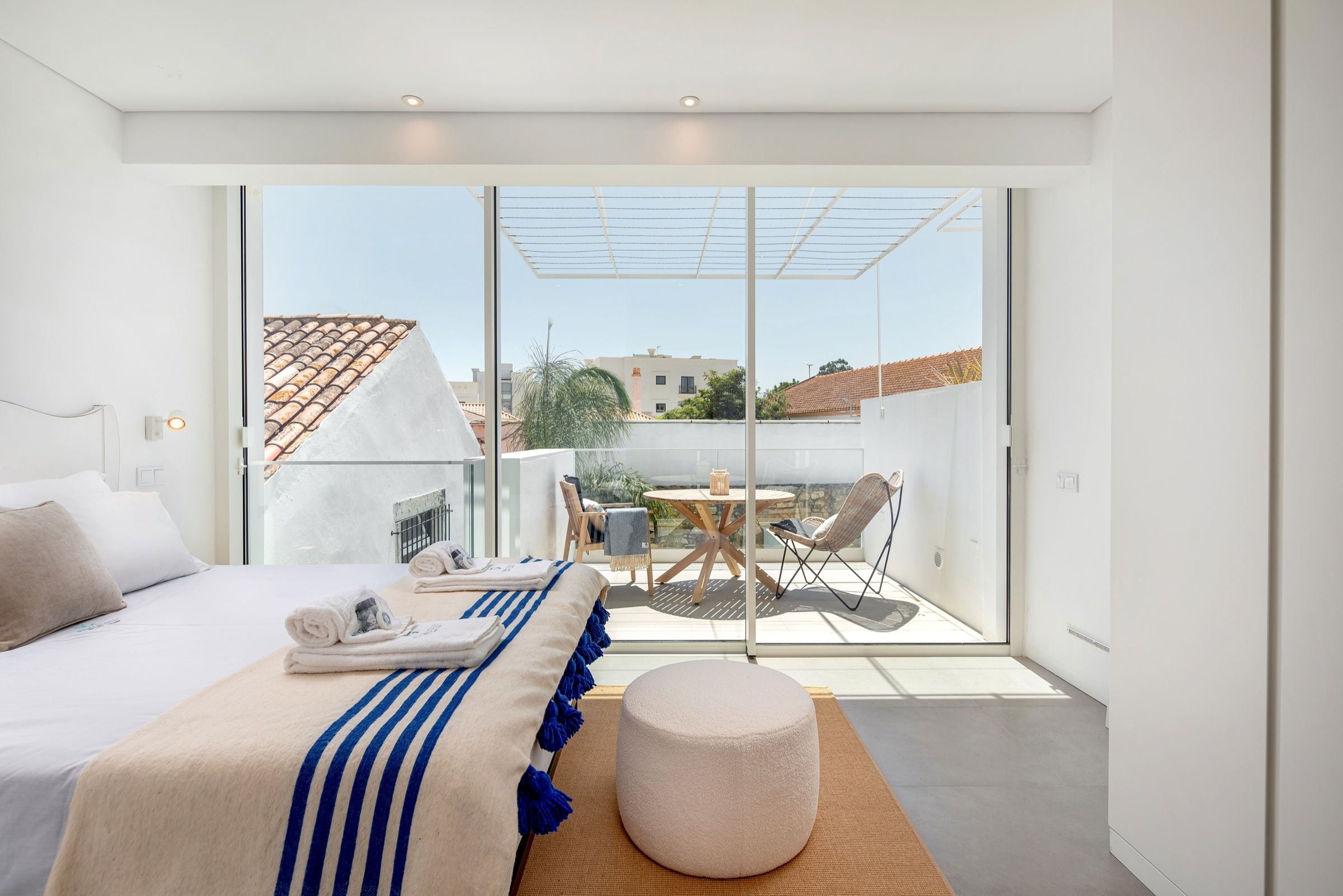 White bedroom with a blue-striped throw and a glass door onto a terrace with a table and chairs
