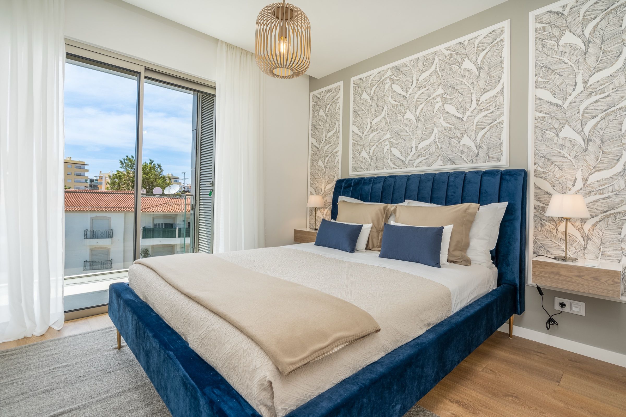 Bedroom with a blue velvet bed, leaf-patterned wallpaper and a full-height window