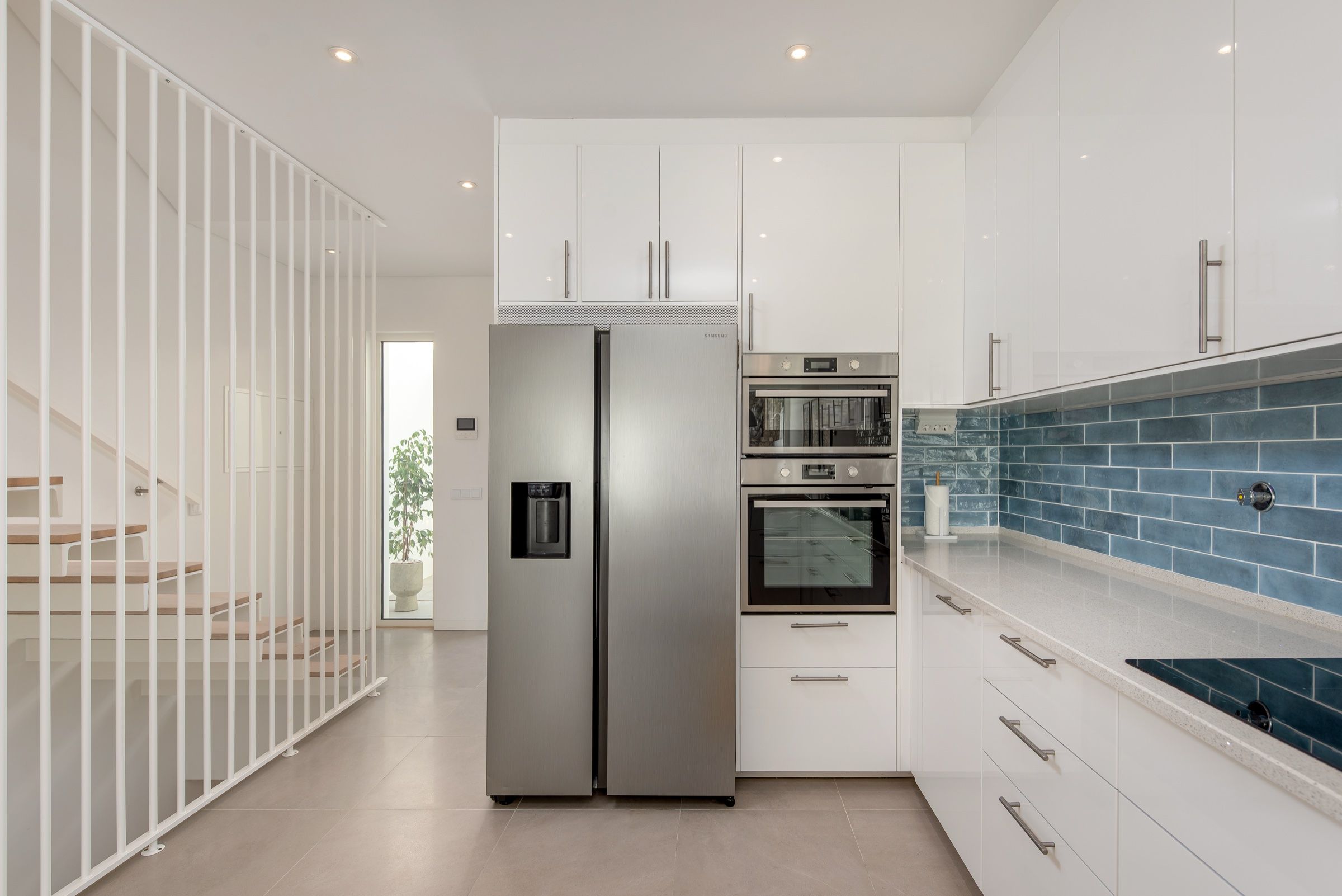 White kitchen with blue tiles, an American-style fridge and a staircase with a white slatted rail