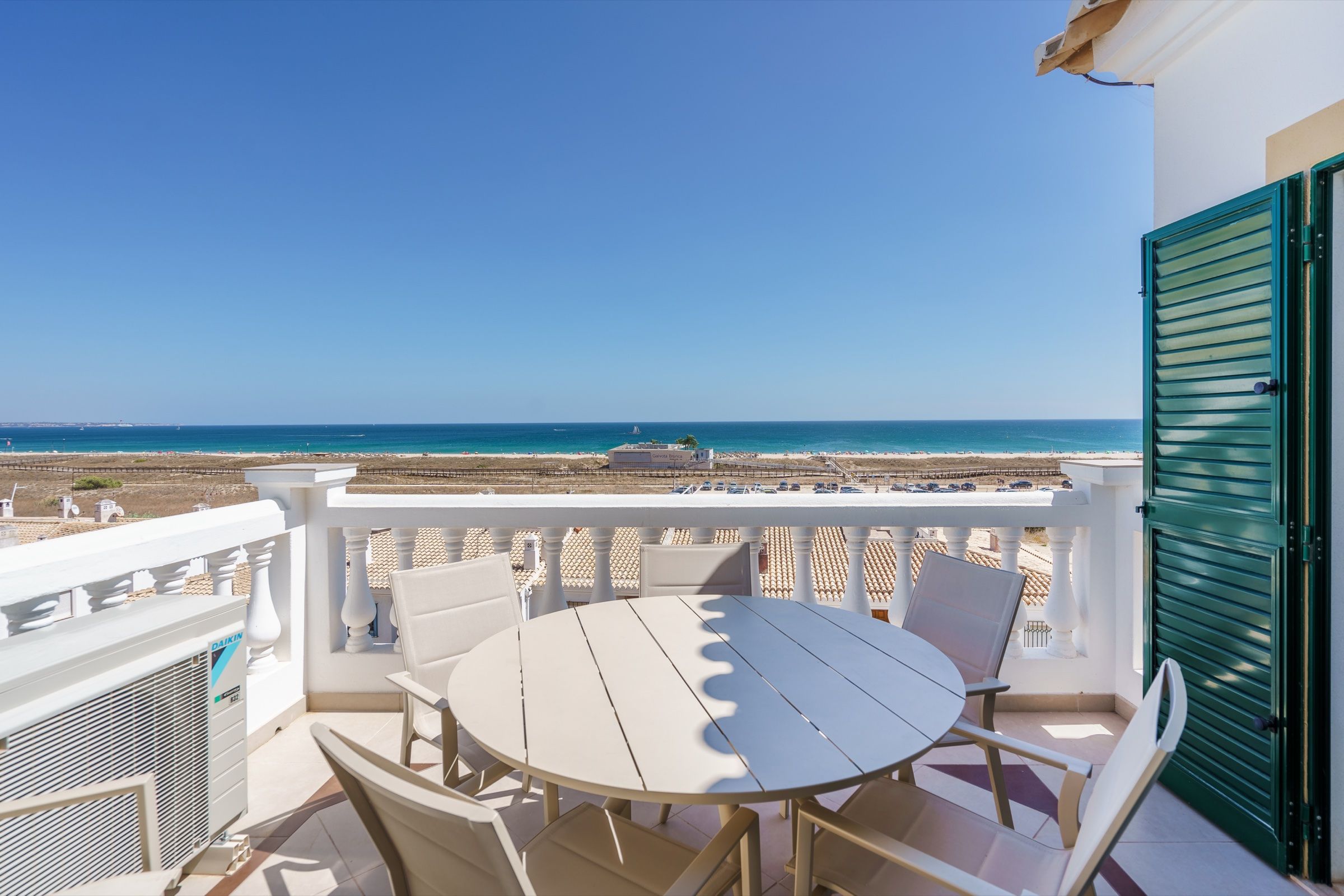 Balcony with a round table and chairs, a white balustrade and green shutters, facing the beach and the sea