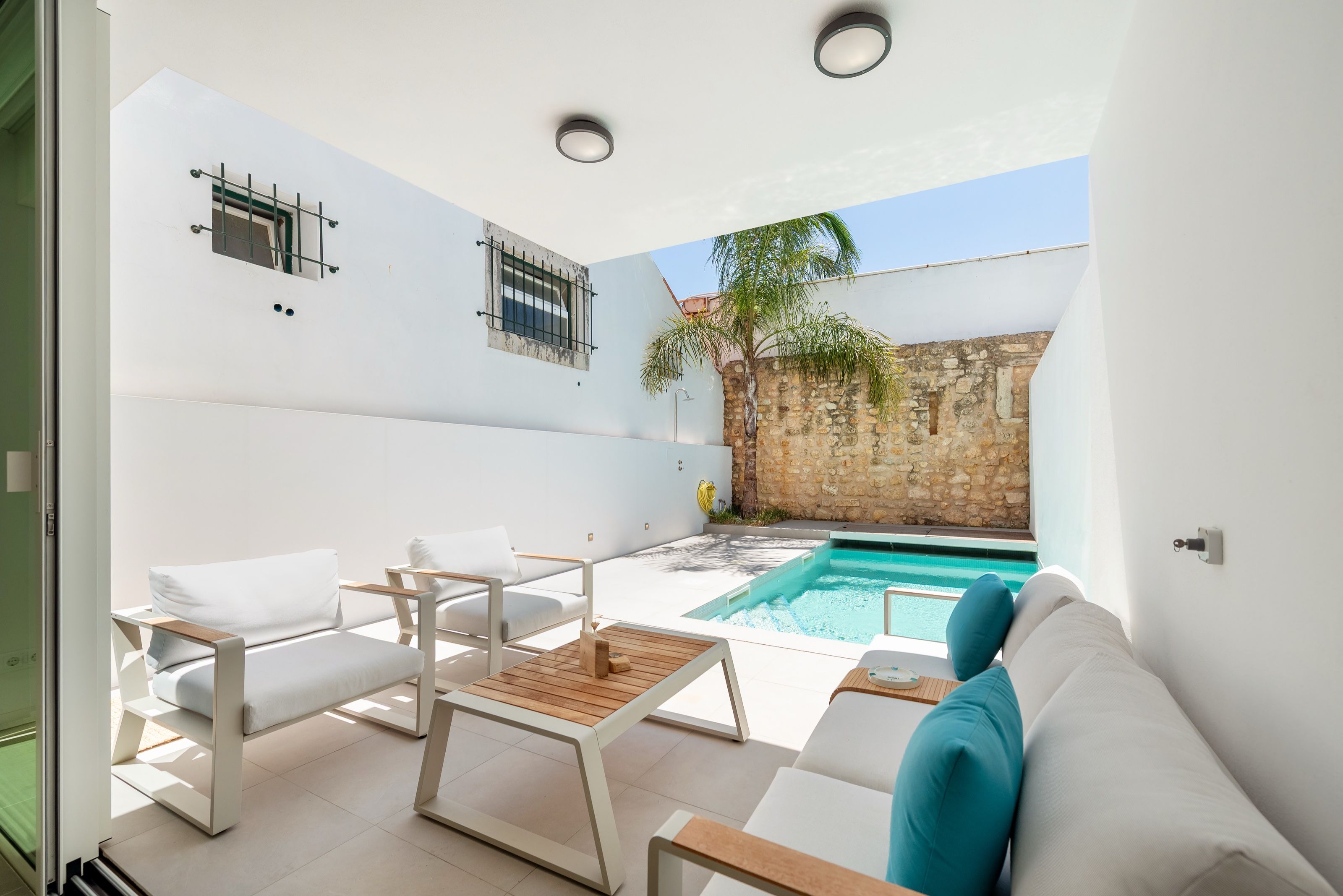 Covered patio of a white house with outdoor sofas, opening onto a pool with a palm tree and a stone wall