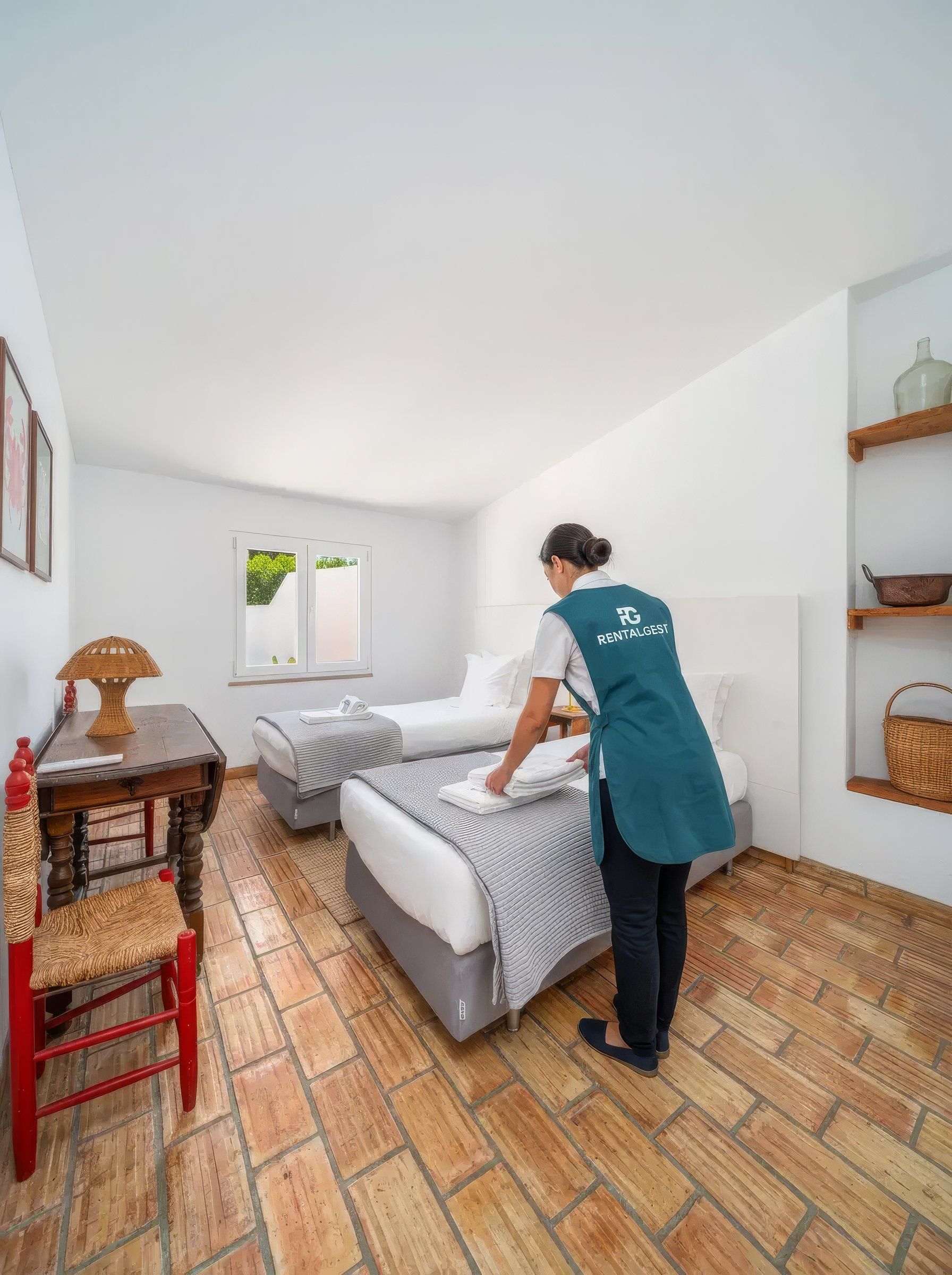 Housekeeper in a Rentalgest apron placing folded towels on one of the beds in a twin room with a terracotta tiled floor